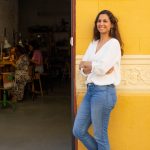Ganzkörperportrait einer Goldschmiedin vor ihrer Werkstatt während innen das freie Atelier stattfindet und Teilnehmerinnen an ihrem Schmuck arbeiten