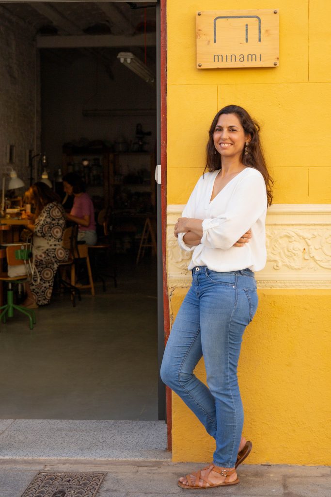 Ganzkörperportrait einer Goldschmiedin vor ihrer Werkstatt während innen das freie Atelier stattfindet und Teilnehmerinnen an ihrem Schmuck arbeiten