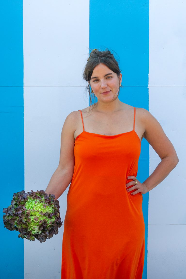 Köchin mit Blick in die Kamera, steht in schickem roten Kleid draußen vor einer blau/weissen Wand und hat einen Salat in der Hand