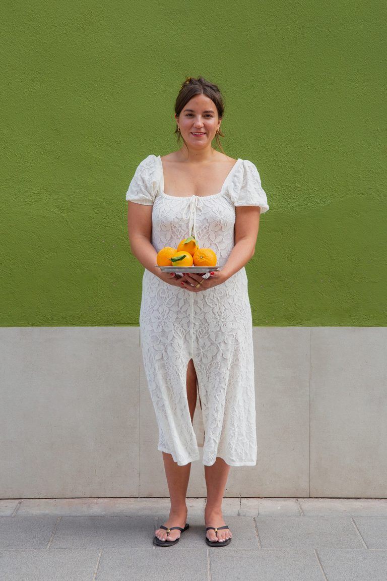 Köchin mit weißem Kleid und Blick in die Kamera steht draußen vor einer Wand mit einer Orangenschale in den Händen